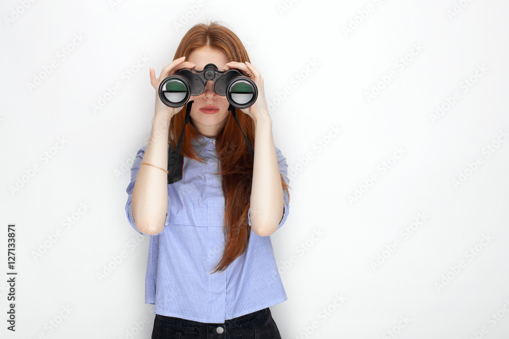 Portrait of cute redhead girl wearing blue striped shirt smiling with happiness and joy looking thru binoculars while posing against white studio background Portrait of cute redhead girl wearing blue striped shirt smiling with happiness and joy looking thru binoculars while posing against white studio background