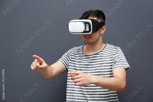 Young man in striped T-shirt wearing virtual reality glasses on grey background