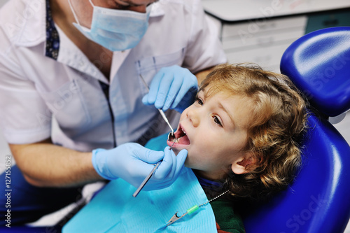 Male dentist examines the teeth of the patient cheerful child with curly red hair. Moloi boy smiling in dentist s chair. child mouth wide open in the dentist s chair