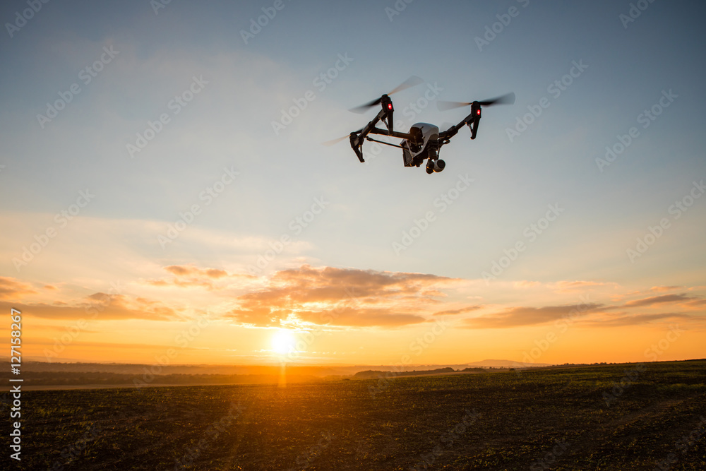 White drone with digital camera flying in sky over field on sunset White drone with digital camera flying in sky over field on sunset