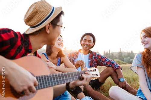 Happy young people sitting outdoors and playing guitar together