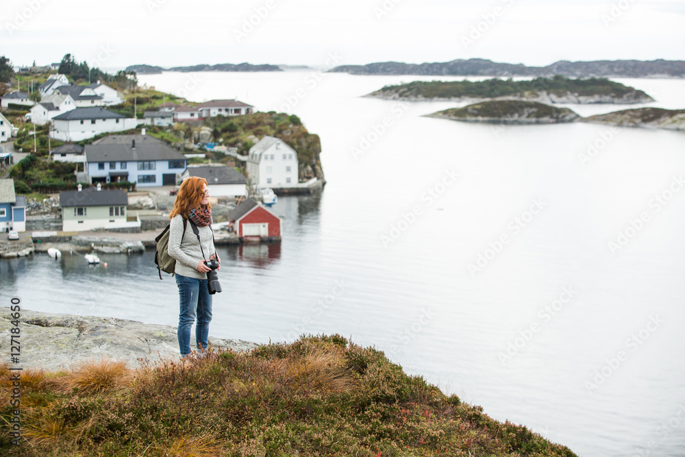 Red-haired girl with glasses standing on top of a hill on a background of ocean and islands. Overcast. Scandinavia.