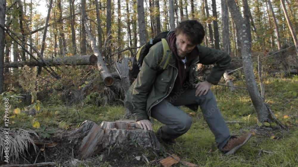 A middle aged man with a beard wearing grey pants, green jacket and yellow boots going uphill sitting on a tree stub to take breath while looking around. A middle aged man with a beard wearing grey pants, green jacket and yellow boots going uphill sitting on a tree stub to take breath while looking around.