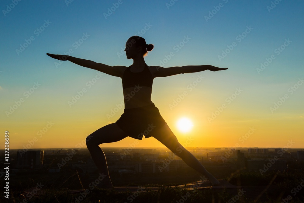 Silhouette of sporty woman doing Ashtanga yoga in the park at sunset. Sunset light, sun lens flares, golden hour. Freedom, health and yoga concept