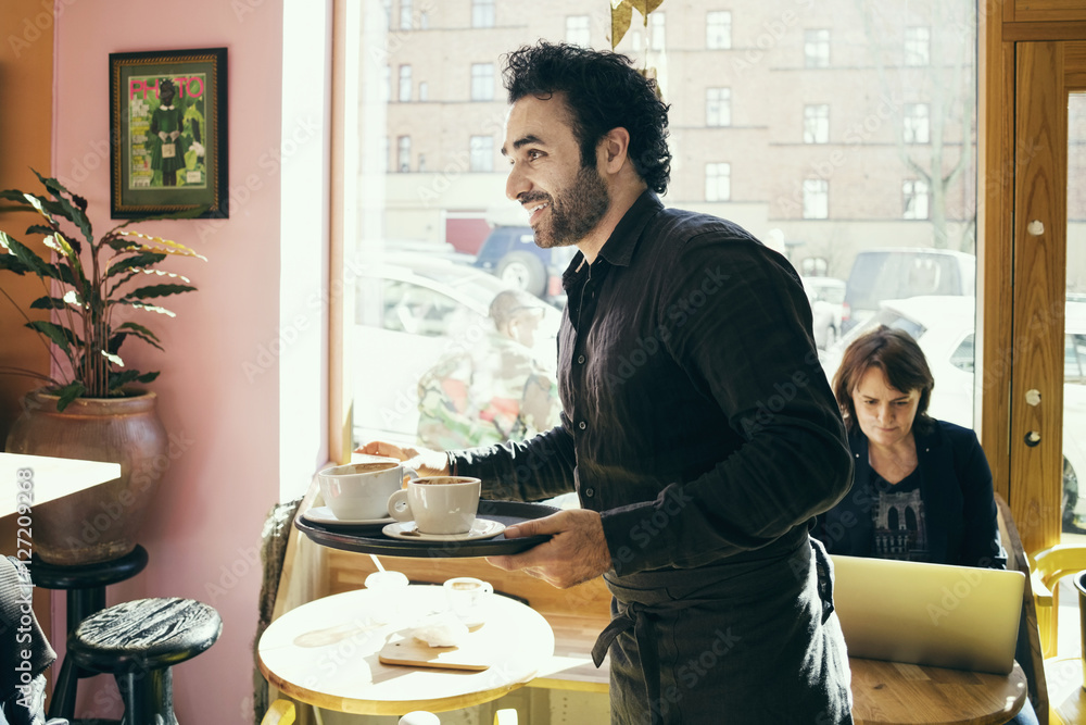 Smiling waiter holding serving tray against customer using laptop in background at cafe Smiling waiter holding serving tray against customer using laptop in background at cafe