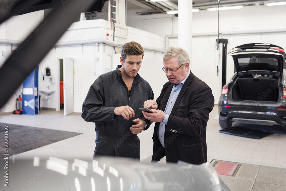 Male customer showing mobile phone to mechanic at auto repair shop Male customer showing mobile phone to mechanic at auto repair shop