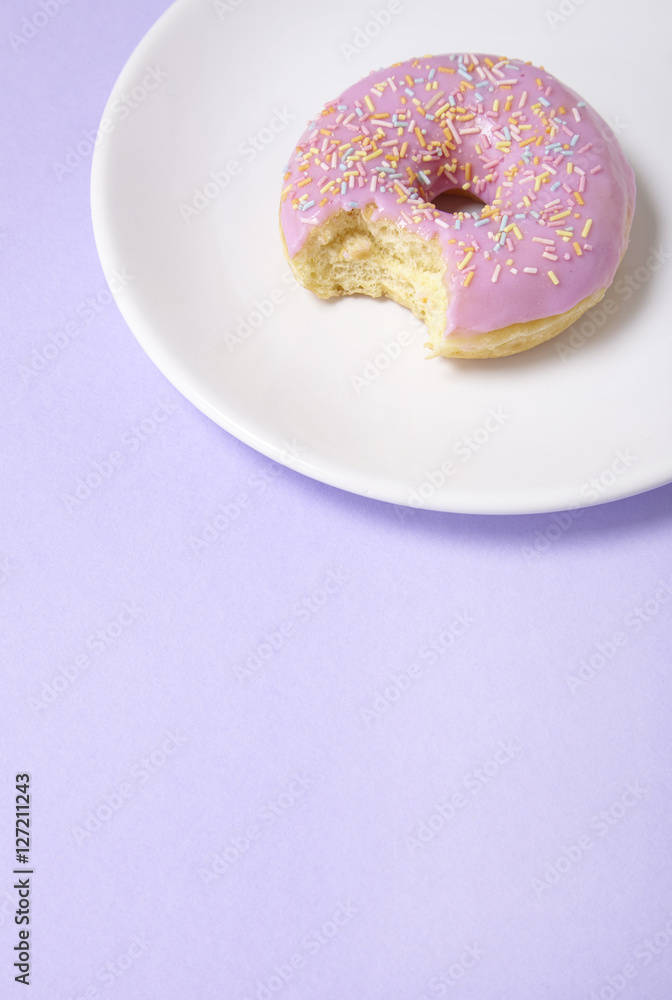A ring doughnut with pastel pink frosting and sprinkles on a purple background, with a bite missing and blank space below A ring doughnut with pastel pink frosting and sprinkles on a purple background, with a bite missing and blank space below