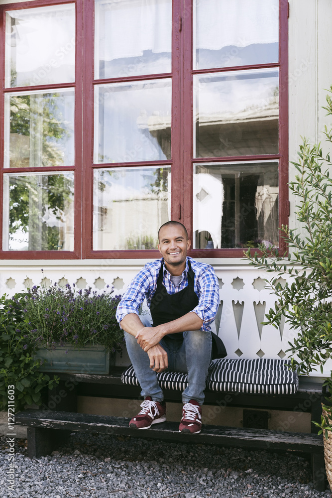 Portrait of smiling male owner resting on bench against window Portrait of smiling male owner resting on bench against window