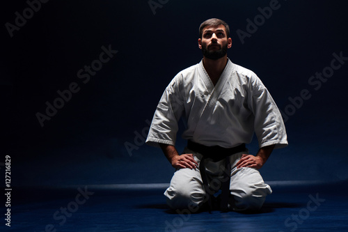 Karate man in a kimono sits on her knees on the floor on blue background