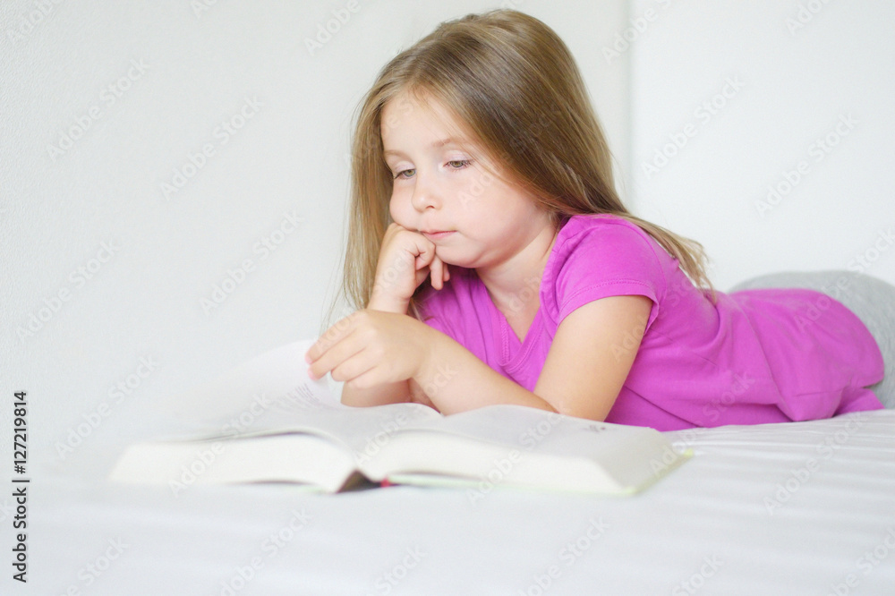 Adorable little girl lying on the bed and reading a book