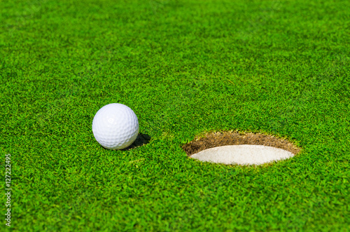 A hole over golf ball on green. Shallow depth of field. Focus on the ball and the hole.
