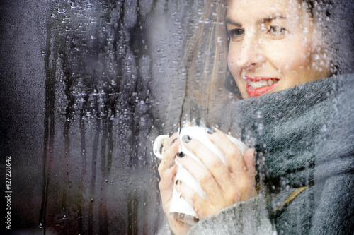 attractive adult woman drinking coffee and looking out of the window on rainy day, close up portre in winter time