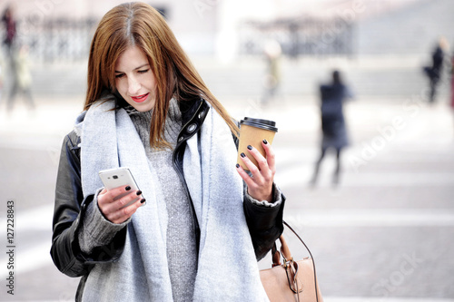 Fashionable young hipster woman in trendy oufit is looking her mobile with a smile on her face. City life background. photo