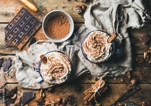 Hot chocolate with whipped cream and cinnamon sticks served with anise, nuts and cocoa powder on rustic wooden background, top view, horizontal composition