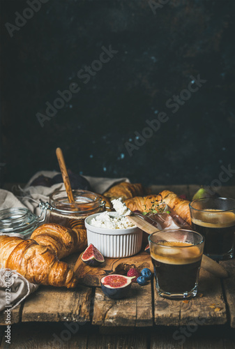 Breakfast with croissants, ricotta cheese, figs, fresh berries, prosciutto meat, honey and espresso coffee on rustic wooden table, dark blue plywood wall background, selective focus, copy space