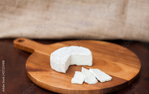 Brie or camembert cheese cut out wedge and in slices on a wooden chopping cutting cheese board on dark brown wooden table with a light brown burlap background