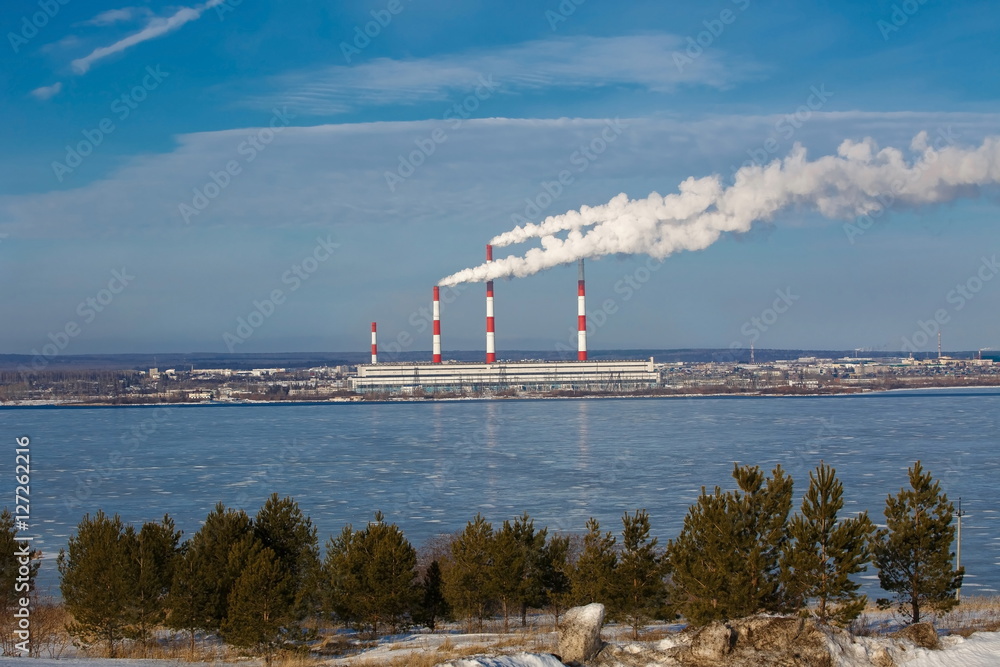 Industrial landscape with the steam from the pipes thermoelectric power station on a background of blue sky and the river in winter