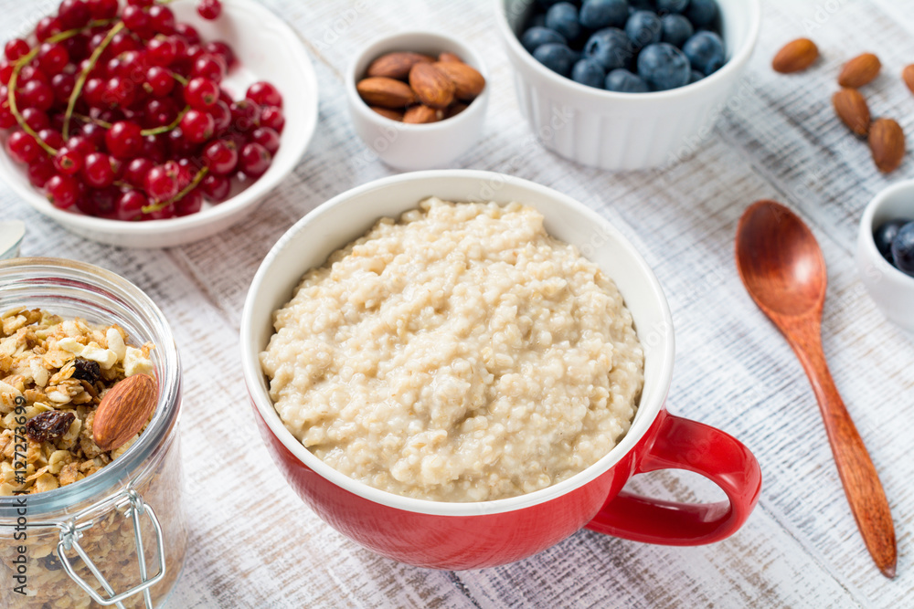 Scottish porridge oats in red ceramic bowl. Served with red currants, fresh blueberries, almonds and muesli. White table background. Healthy vegan eating concept, bright image of healthy breakfast Scottish porridge oats in red ceramic bowl. Served with red currants, fresh blueberries, almonds and muesli. White table background. Healthy vegan eating concept, bright image of healthy breakfast