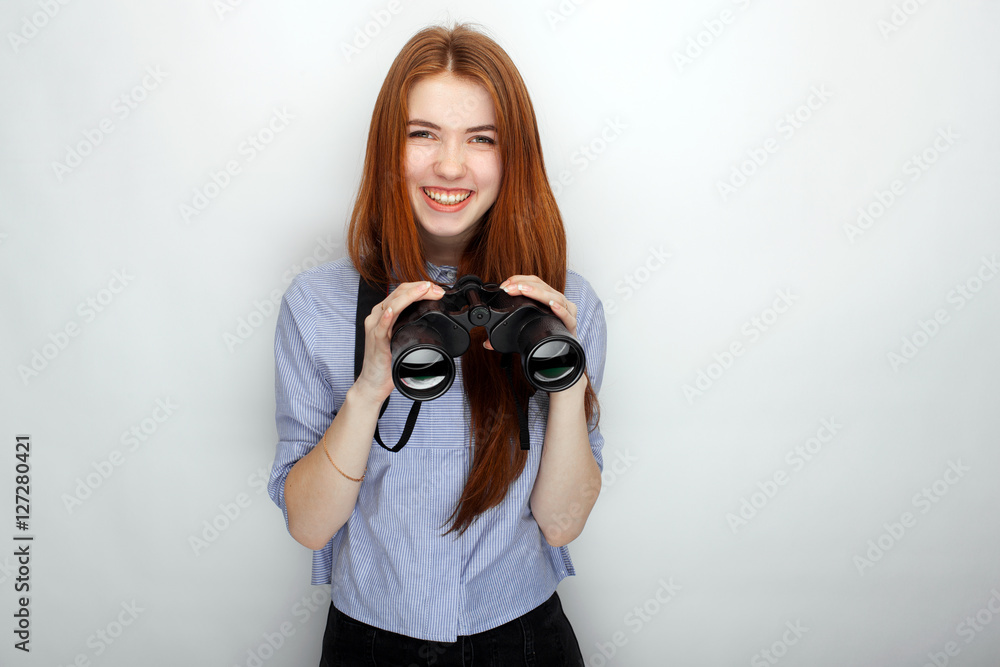 Portrait of young cute redhead woman  wearing blue striped shirt smiling with happiness and joy while posing with binoculars against white studio background.