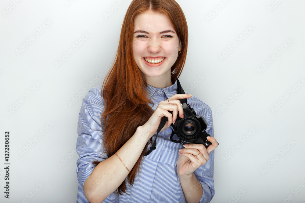 Portrait of cute redhead photographer woman wearing blue striped shirt smiling with happiness and joy while posing with camera against white studio background. Portrait of cute redhead photographer woman wearing blue striped shirt smiling with happiness and joy while posing with camera against white studio background.