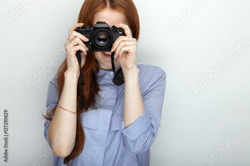 Portrait of cute redhead photographer woman wearing blue striped shirt smiling with happiness and joy while posing with camera against white studio background.
