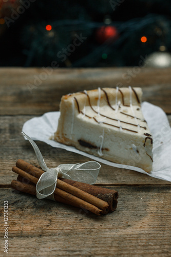 Beautiful homemade cake cheesecake on a white paper on the wooden background with cinnamon stick. Near Christmas gifts, Christmas tree and toys. Conceived to celebrate the new year.
