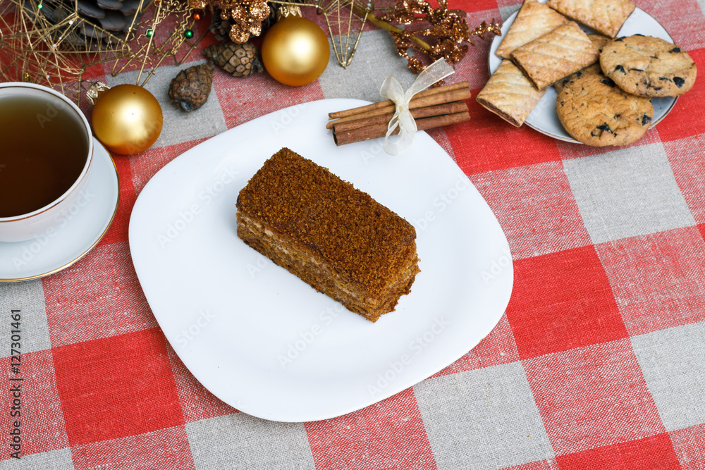 Cake on a white plate on a checkered tablecloth with tea, biscuits, cinnamon on a background of a New Year's Christmas tree. New concept. For holiday. View from above.
