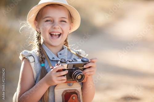 Girl 6 years old with an old brown suitcase and an old camera in hand,brunette with two braids,wearing a straw hat  in a blue flight suit,nature photographs while traveling,standing on a country road