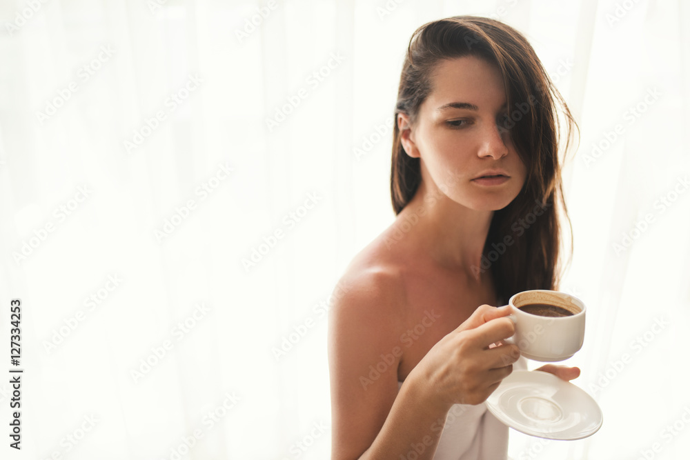 Cute young woman relaxing at cozy atmosphere next to window. Caucasian woman wearing white towel with cup of hot chocolate or coffee in her hands, look at one side. Soft light and natural beauty. Cute young woman relaxing at cozy atmosphere next to window. Caucasian woman wearing white towel with cup of hot chocolate or coffee in her hands, look at one side. Soft light and natural beauty.