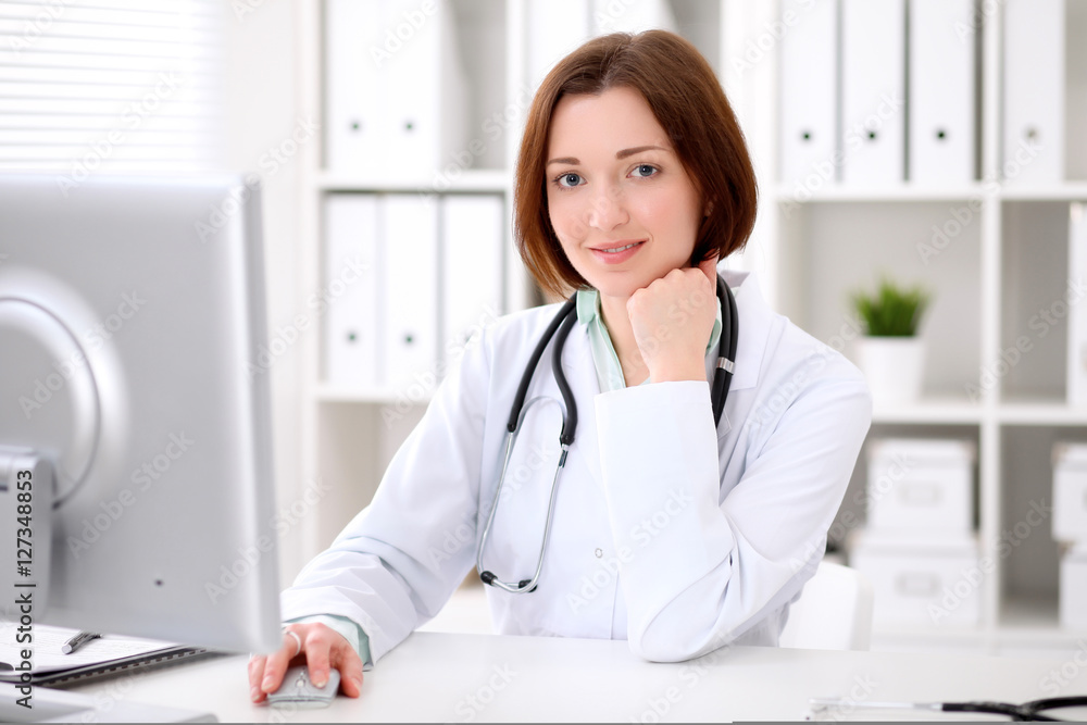 Young brunette female doctor sitting at a desk and working on the computer at the hospital office. Health care, insurance and help concept. Physician ready to examine patient Young brunette female doctor sitting at a desk and working on the computer at the hospital office. Health care, insurance and help concept. Physician ready to examine patient