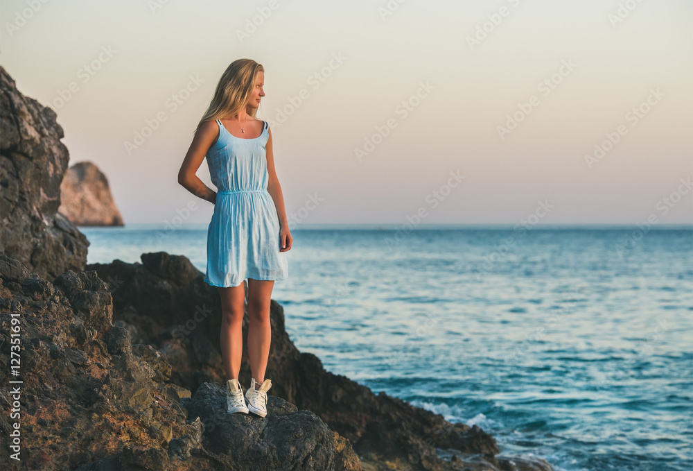 Young blond tourist woman in blue dress standing on natural rocks by the sea at sunset and looking aside. Kleopatra beach, Alanya, Mediterranean region, Turkey Young blond tourist woman in blue dress standing on natural rocks by the sea at sunset and looking aside. Kleopatra beach, Alanya, Mediterranean region, Turkey