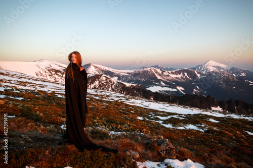 Young woman in long mystical dress looking into the sun stands on the top of the mountain. Woman on the background of snow winter and mounts