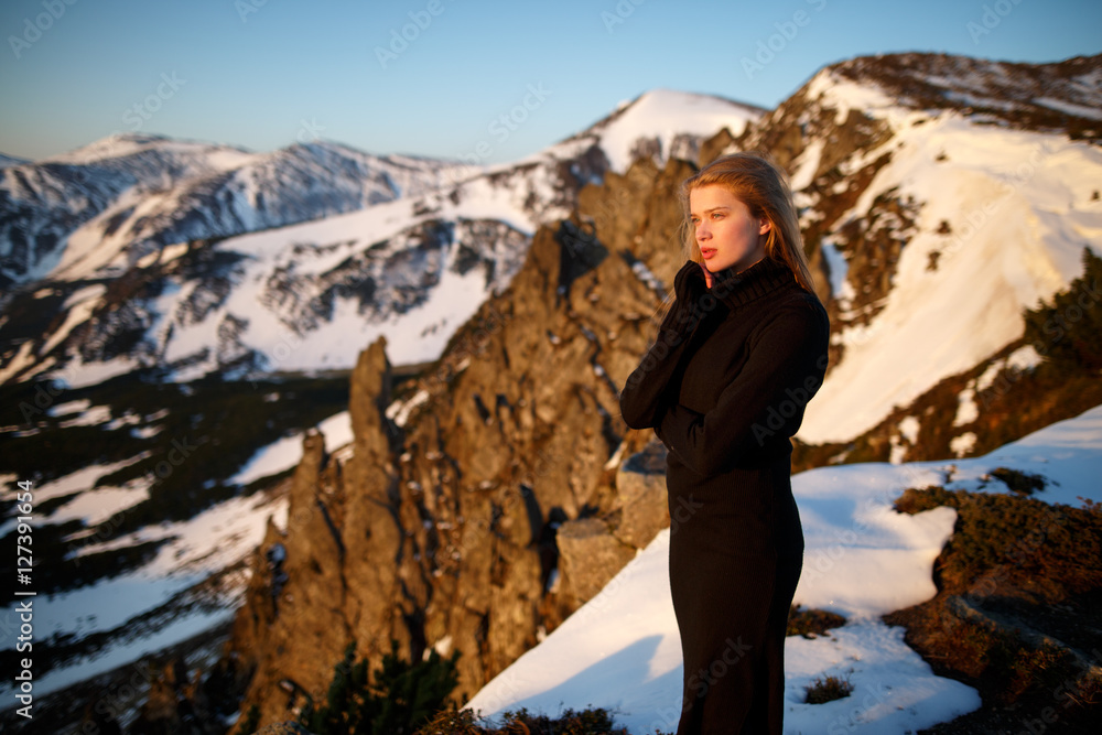 Young woman in long mystical dress looking into the sun stands on the top of the mountain. Woman on the background of snow winter and mounts. Travel inspiration and motivation, beautiful landscape.