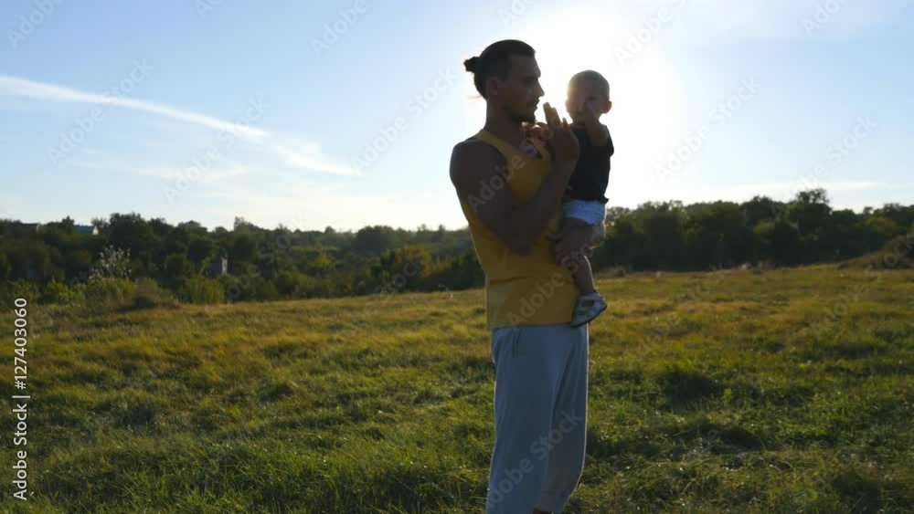 Young father holds on hands his son in the meadow at nature at sunny summer day. Dad and little boy hugging outdoor and giving five to each other. Beautiful landscape at background. Close up Young father holds on hands his son in the meadow at nature at sunny summer day. Dad and little boy hugging outdoor and giving five to each other. Beautiful landscape at background. Close up