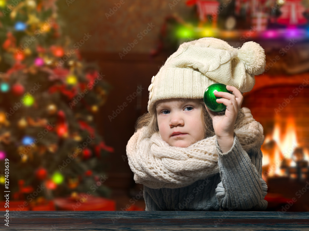 Serious little girl shows a green Christmas ball. House with Christmas decor. Christmas tree, fireplace. Child in hat and scarf Serious little girl shows a green Christmas ball. House with Christmas decor. Christmas tree, fireplace. Child in hat and scarf