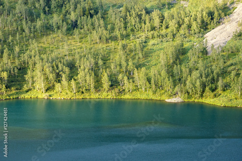 Russia. Khakassia. Sayan Mountains. Reserve Kuznetsky Alatau. Ivanovskie lake. The light of the setting sun behind the mountain on the turquoise water. Riverside alpine lake, wooded.