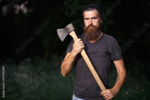 Brutal brunette bearded man in warm hat with a hatchet in the woods on a background of trees