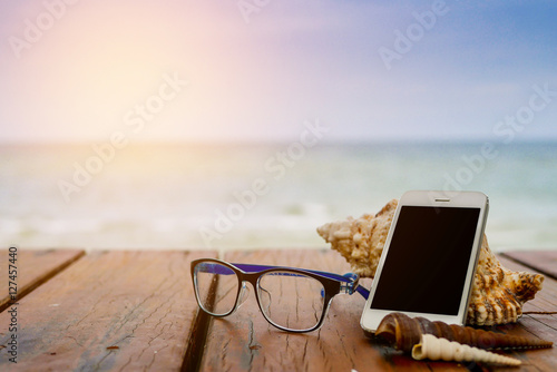 Smartphone and sea shells with glasses on brown wooden table. Ti