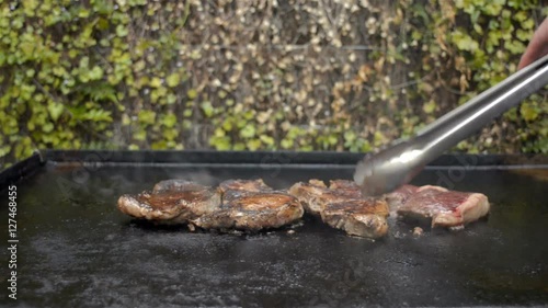 Steaks being cooked on a BBQ, with a hand coming in to turn them over with tongs as they are being cooked.