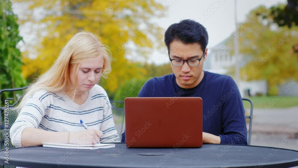 Young students - Asian man and Caucasian woman engaged outdoors. Enjoy a laptop woman takes notes in a notebook Young students - Asian man and Caucasian woman engaged outdoors. Enjoy a laptop woman takes notes in a notebook