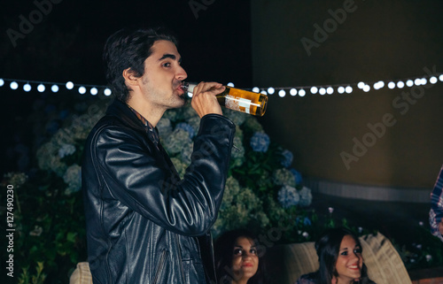 Portrait of young man driking bottle of beer while having fun with his friends in a outdoors party. Friendship and celebrations concept.