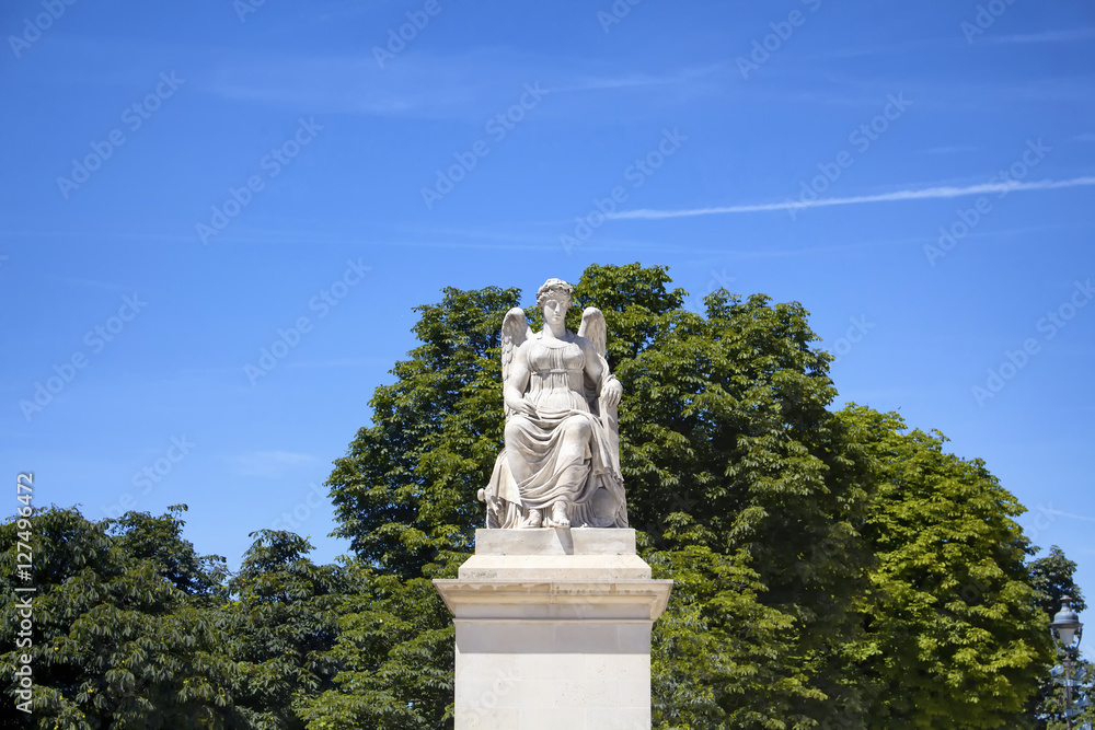 View of winged woman statue at Jardin Des Tuileries in Paris. Trees and blue sky are in the background.