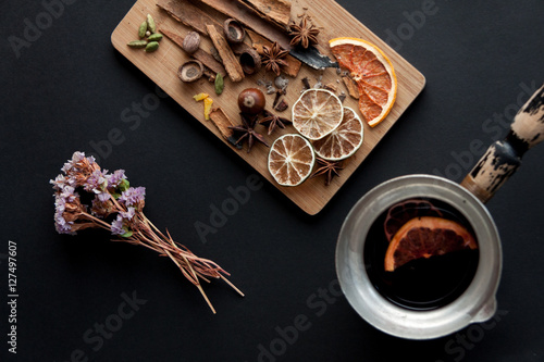 Cutting board with mixed spices and dry citrus slices . On the black background with some desiccated flowers on the left t side and a coffee pot with mulled wine inside . © Regina