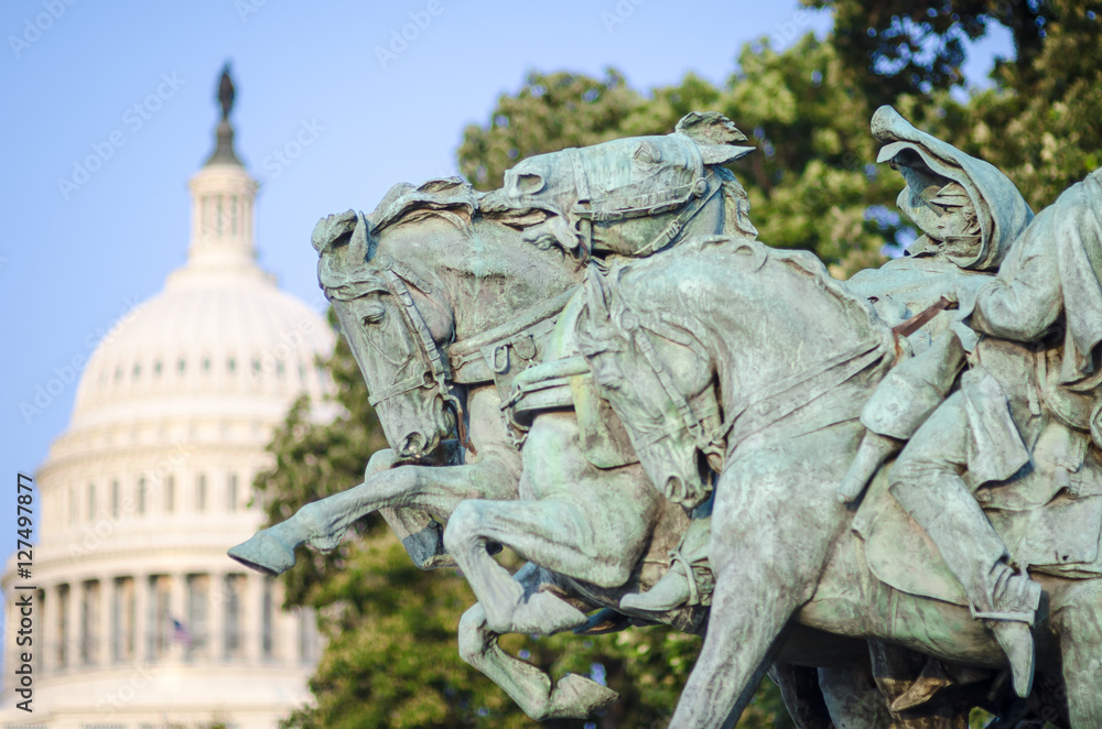 Obraz premium Bronze memorial of horses (Artillery, 1912) charging forward with the US Capitol Building dome under blue sky in the background in Washington, DC