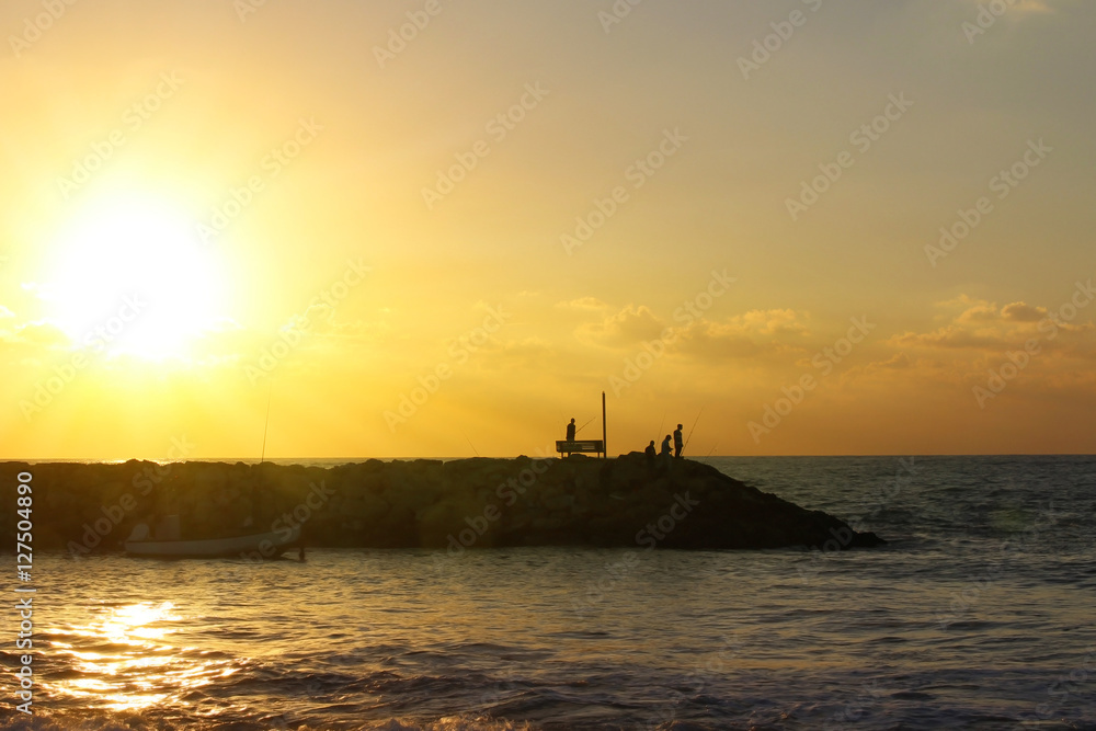 Silhouettes of fishermen at sunset. Rocky shore for fishing. Evening sky and quiet sea background.