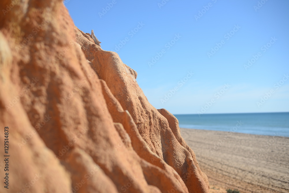 Closeup on red rock wall with defocused beach and sea. Sunny outdoors background