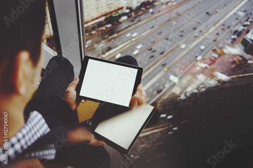 Mock up template of tablet pc in hands of businessman sitting near window of skyscraper and using modern digital tablet with blank screen for your text message, cityscape and highway outside