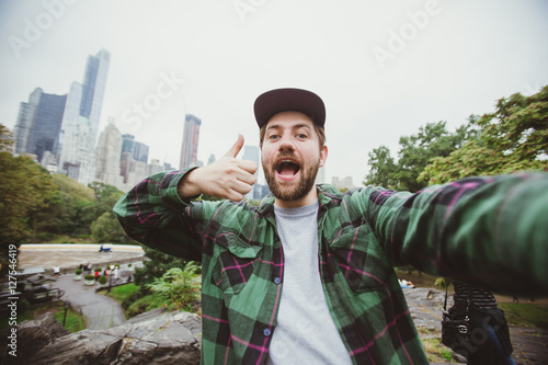 Handsome bearded hipster taking selfie with camera in Central Park, New York. Happy student makes funny picture for his blog.