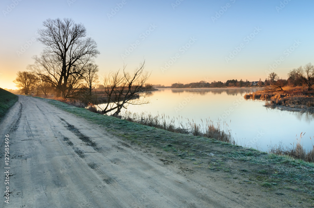 Road on the shore of a wild pond next to a village during sunrise in autumn morning