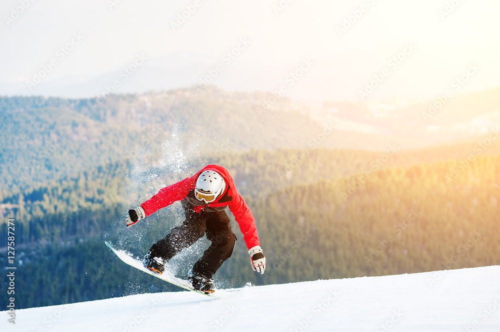 Snowboarder standing on his snowboard and taking his for the edge on top of a mountain against the backdrop of mountains, hills and forests in the distance.